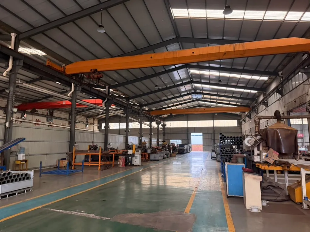 Interior of the workshop hall, with an overhead crane spanning the bay above pipes, steel frames, workbenches and machinery along both sides